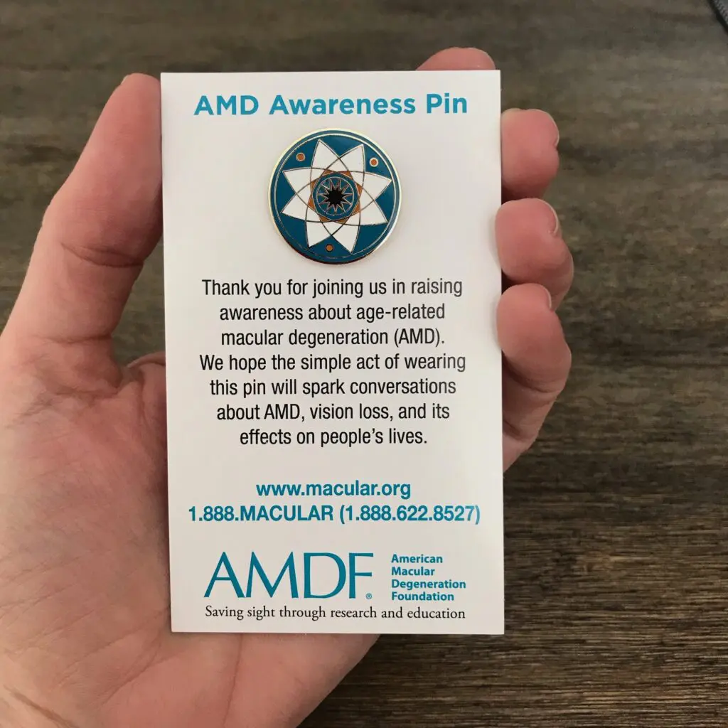 Hand holding card with AMD Awareness Pin attached. Hand holding card with AMD Awareness Pin attached. Text reads: Thank you for joining us in raising awareness about age-related macular degeneration (AMD). We hope the simple act of wearing this pin will spark conversations about AMD, vision loss, and its effects on people's lives.